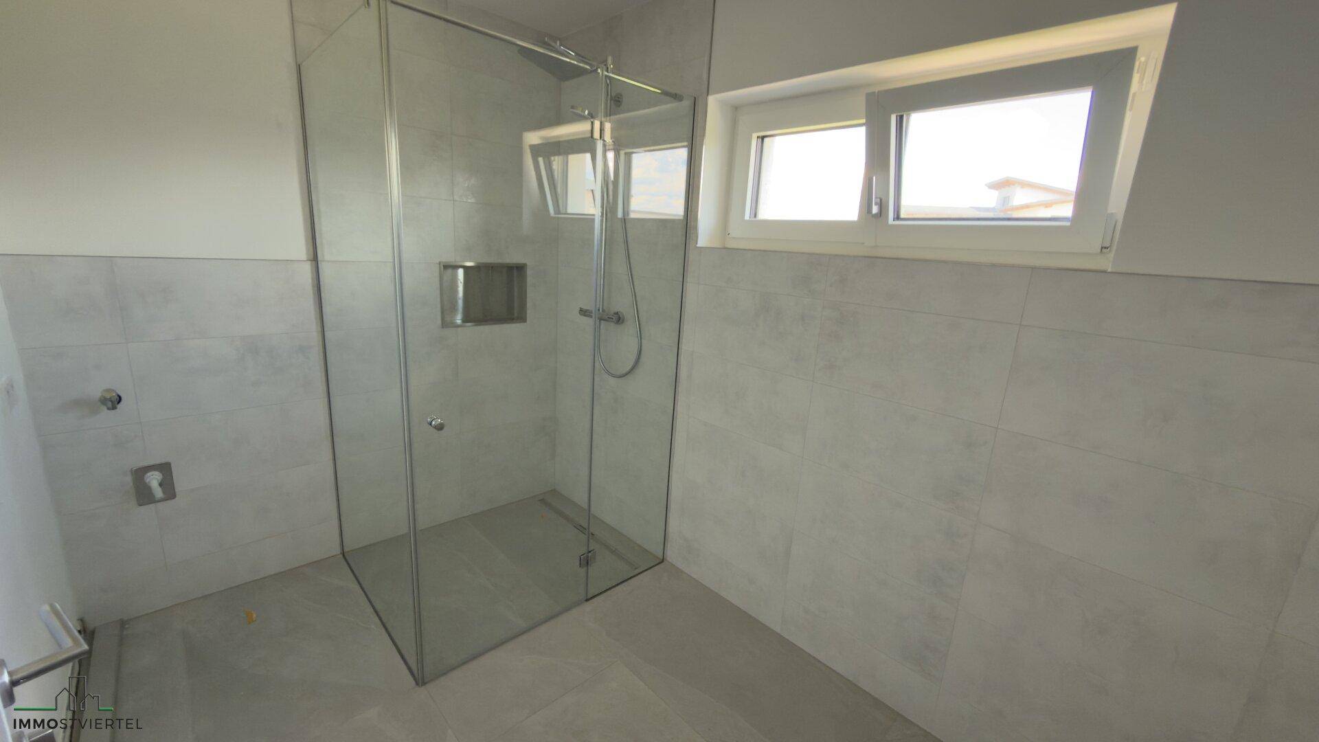Modernes Badezimmer mit ebenerdiger Glasdusche, grauen Fliesen und einem Fenster für Tageslicht.