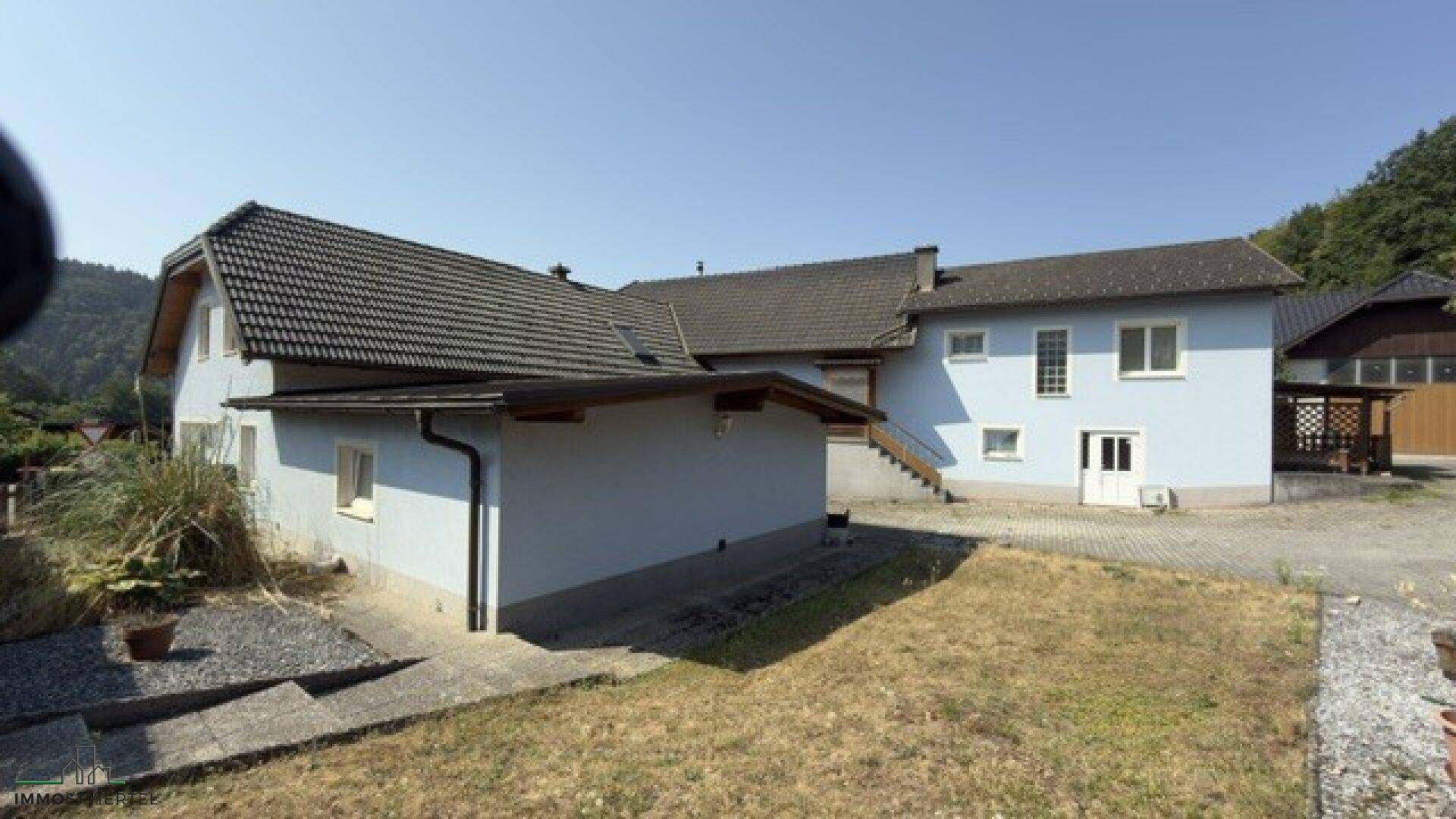 Seitenansicht des Hauses mit hellblauer Fassade, Anbau und einem ungepflegten Gartenbereich.