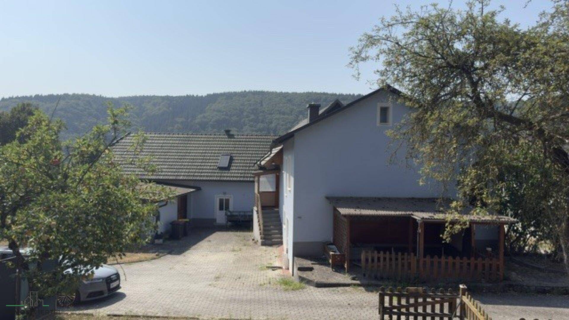 Ansicht des Hauses mit blauer Fassade, überdachtem Bereich und Blick auf bewaldete Hügel.