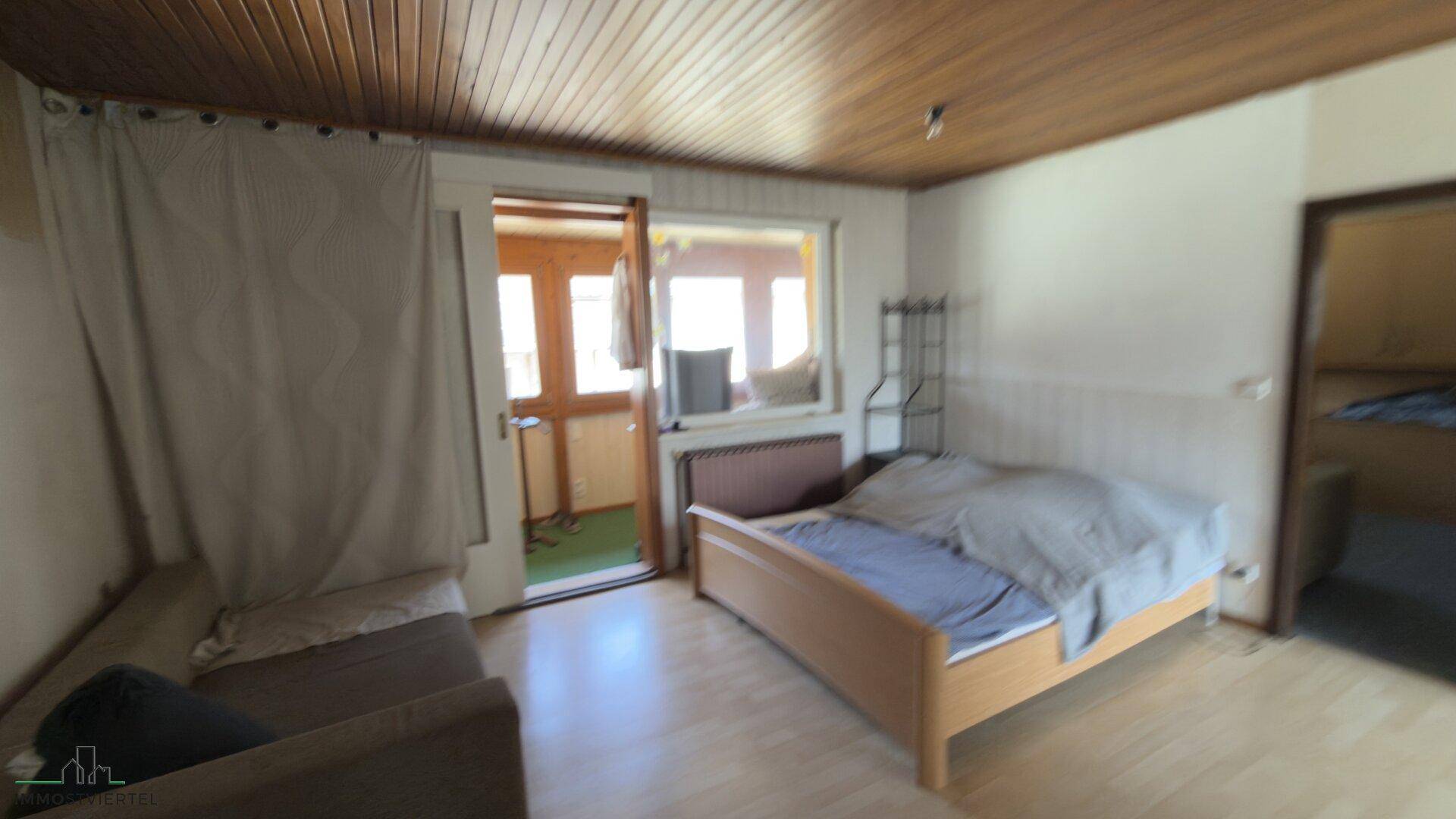 Schlafzimmer mit Holzdecke und hellem Holzboden, Zugang zu einem verglasten Balkon.