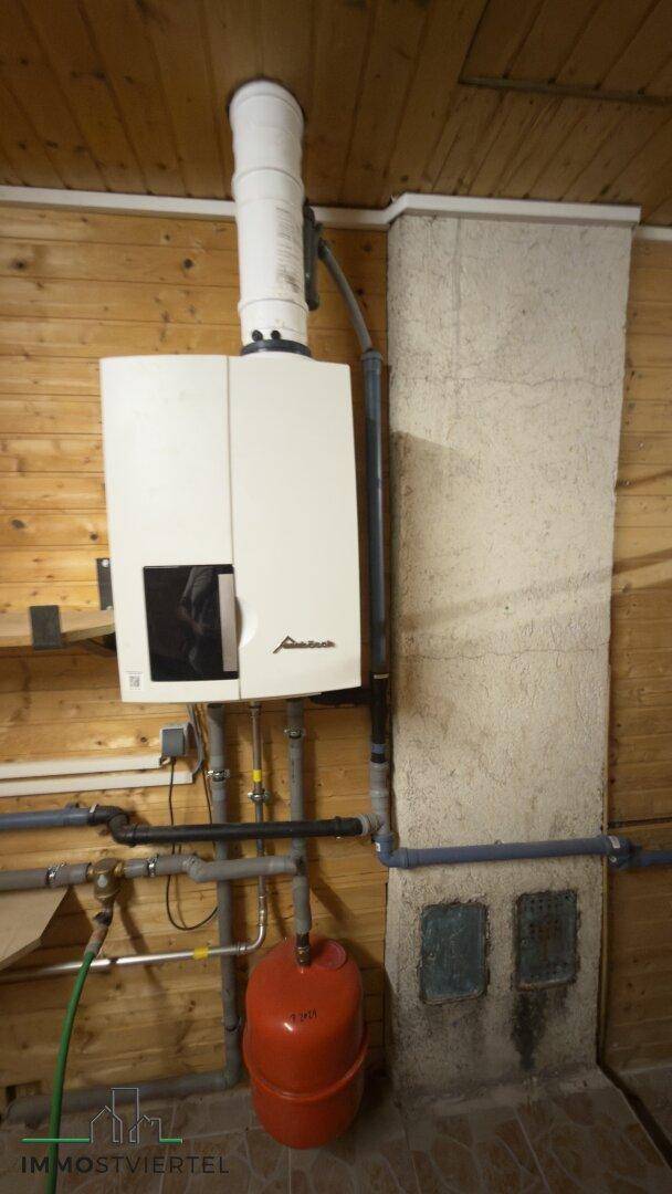 Nahaufnahme der Heizungsanlage mit Rohren und Ausgleichsbehälter in einem Holzraum.
