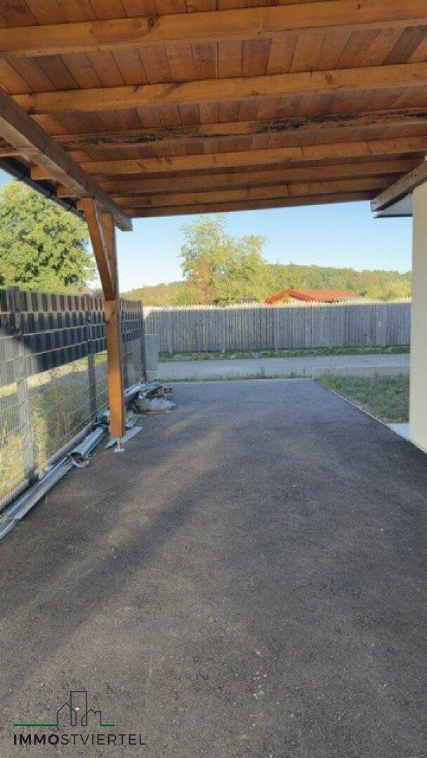Überdachter Stellplatz oder Carport mit Holzbalken und asphaltiertem Boden für Fahrzeuge.