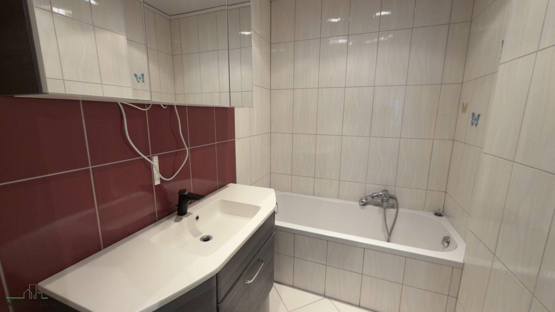 Badezimmer mit Badewanne, Waschtisch und roten Akzentfliesen, funktional eingerichtet.