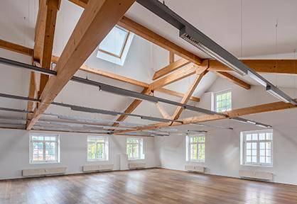 Heller Innenraum mit Holzboden, freiliegenden Holzbalken und mehreren Fenstern, die viel Tageslicht hereinlassen.