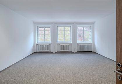 Geräumiger, leerer Raum mit drei Fenstern, Teppichboden und weiß gestrichenen Wänden.