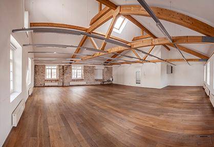 Großer, leerer Raum mit Holzboden, sichtbaren Holzbalken und Dachfenstern, teilweise mit Ziegelwand.
