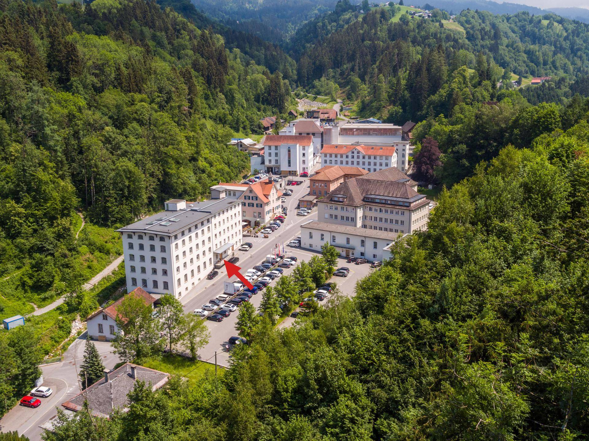 Luftaufnahme eines Gebäudekomplexes in einem grünen Tal, mit einem roten Pfeil, der auf ein Gebäude zeigt.