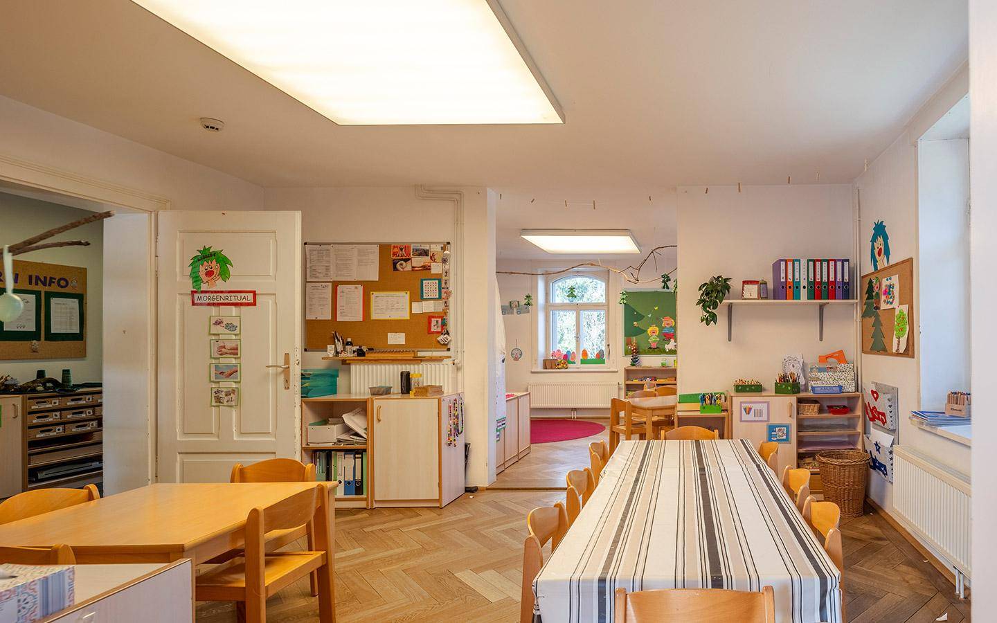 Geräumiger Gruppenraum mit Holzboden und mehreren Tischen, ideal für Lern- und Spielaktivitäten.