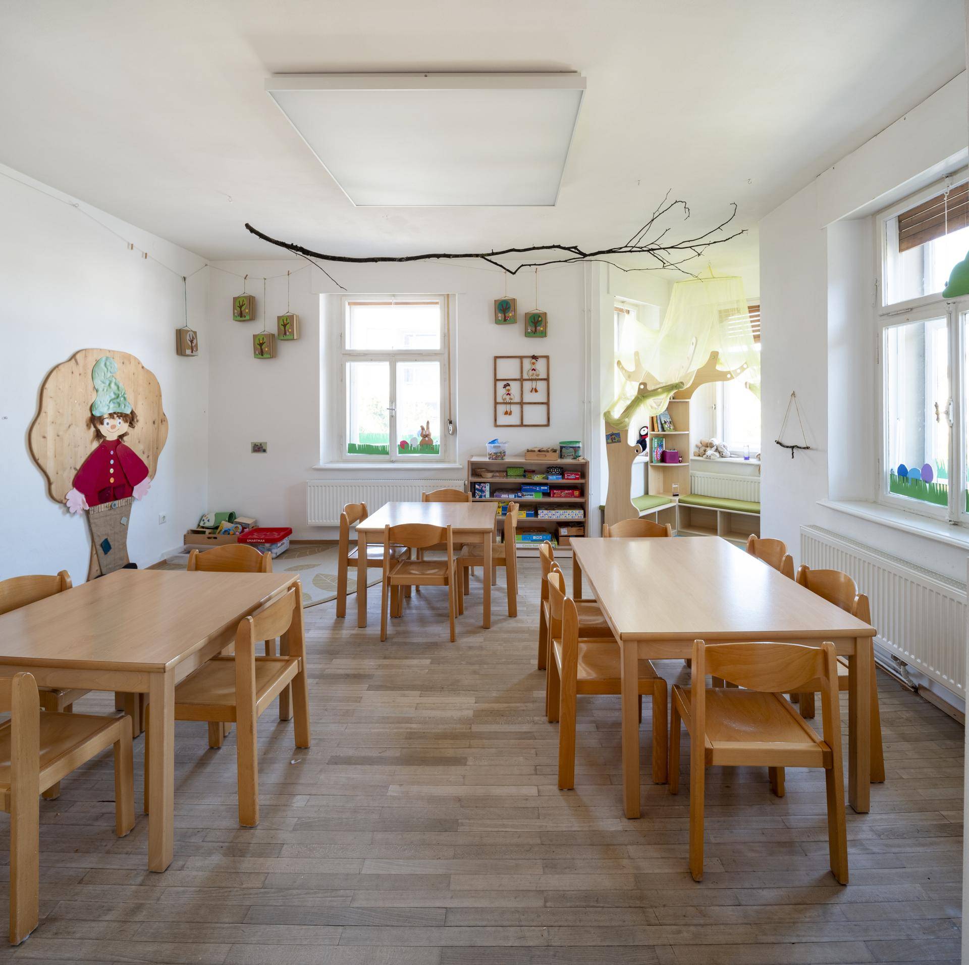 Heller Gruppenraum mit Holztischen und Stühlen, ausgestattet für Kinderaktivitäten und mit Holzparkett.