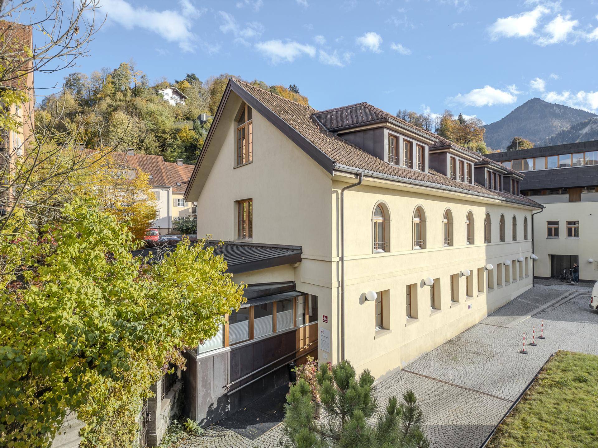 Seitenansicht eines traditionellen Gebäudes mit hellgelber Fassade, Rundbogenfenstern und umgebender Natur.
