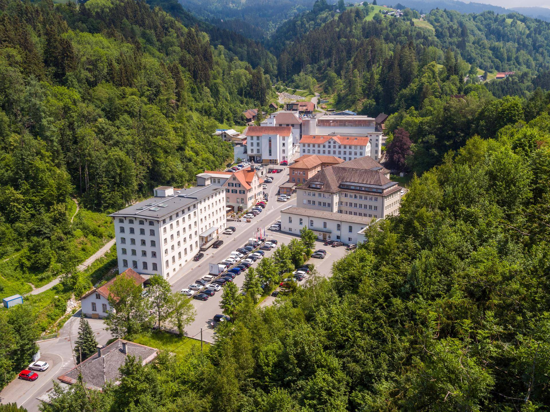 Luftaufnahme eines Gebäudekomplexes in einem grünen Tal, umgeben von dichten Wäldern und Bergen.