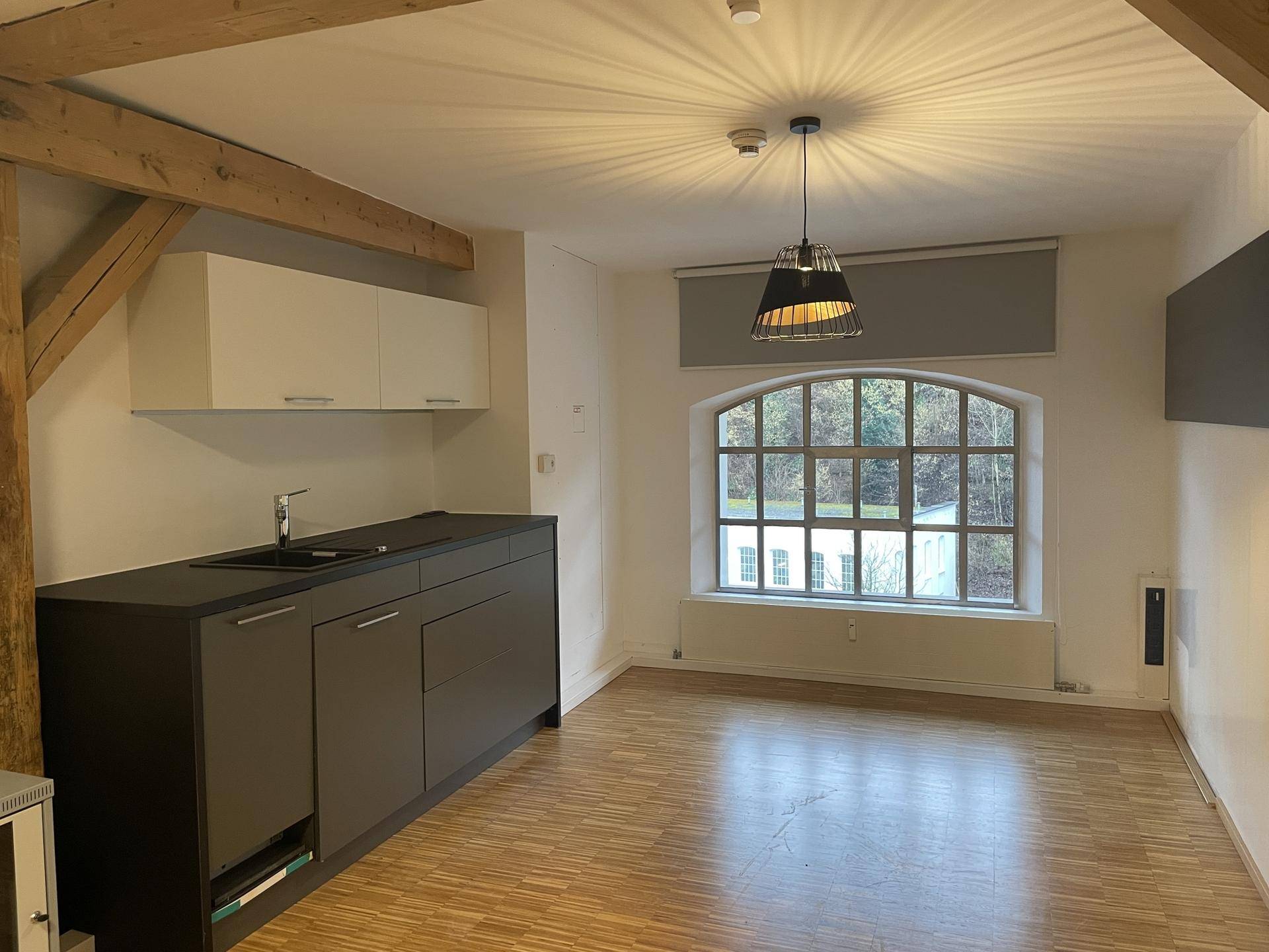 Wohnbereich mit kleiner Küchenzeile, Holzboden, sichtbaren Holzträgern und einem großen Bogenfenster.
