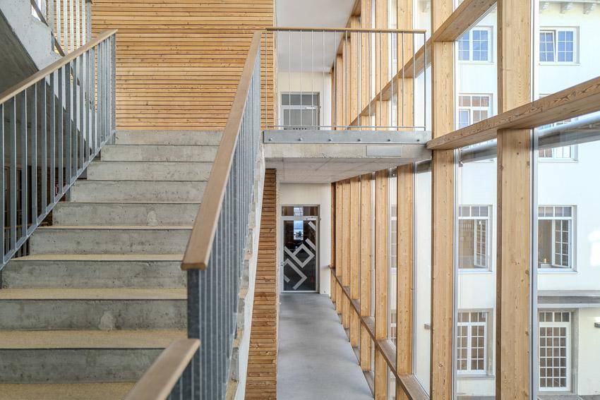 Modernes Treppenhaus mit Betonstufen, Holzverkleidung und großen Fenstern, die viel Licht hereinlassen.