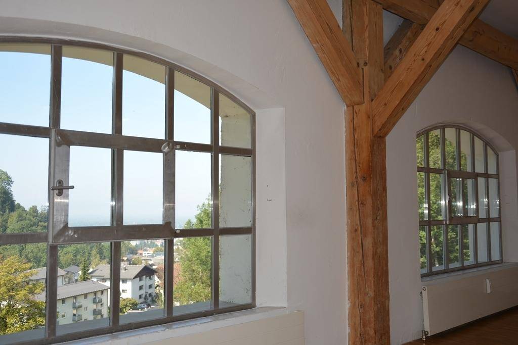 Innenraum mit großen Bogenfenstern, die einen Blick auf die Stadt bieten, und sichtbaren Holzträgern.