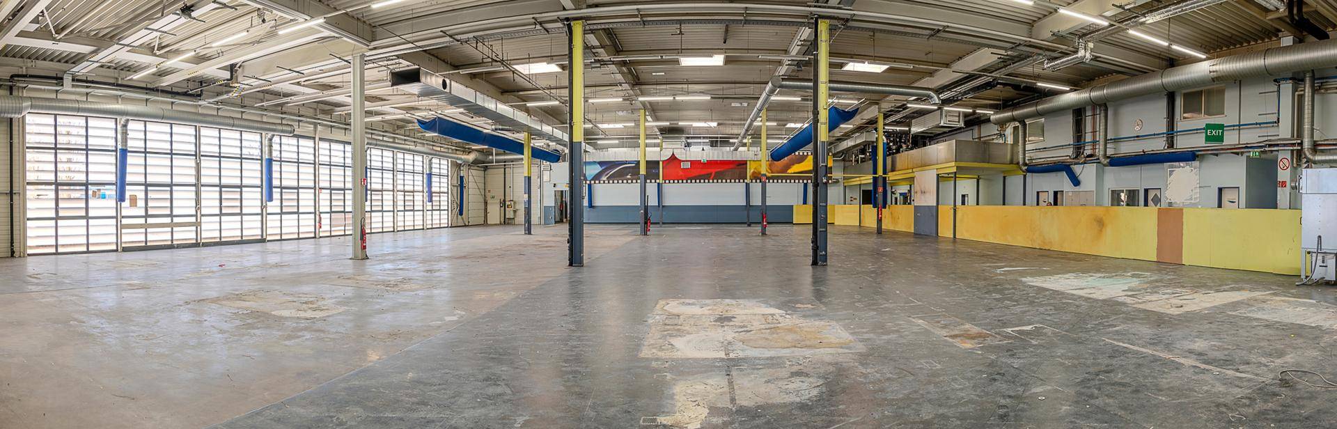 Weitläufige Industriehalle mit Betonboden, sichtbaren Lüftungsrohren und gelben Stützpfeilern, die viel Raum bietet.