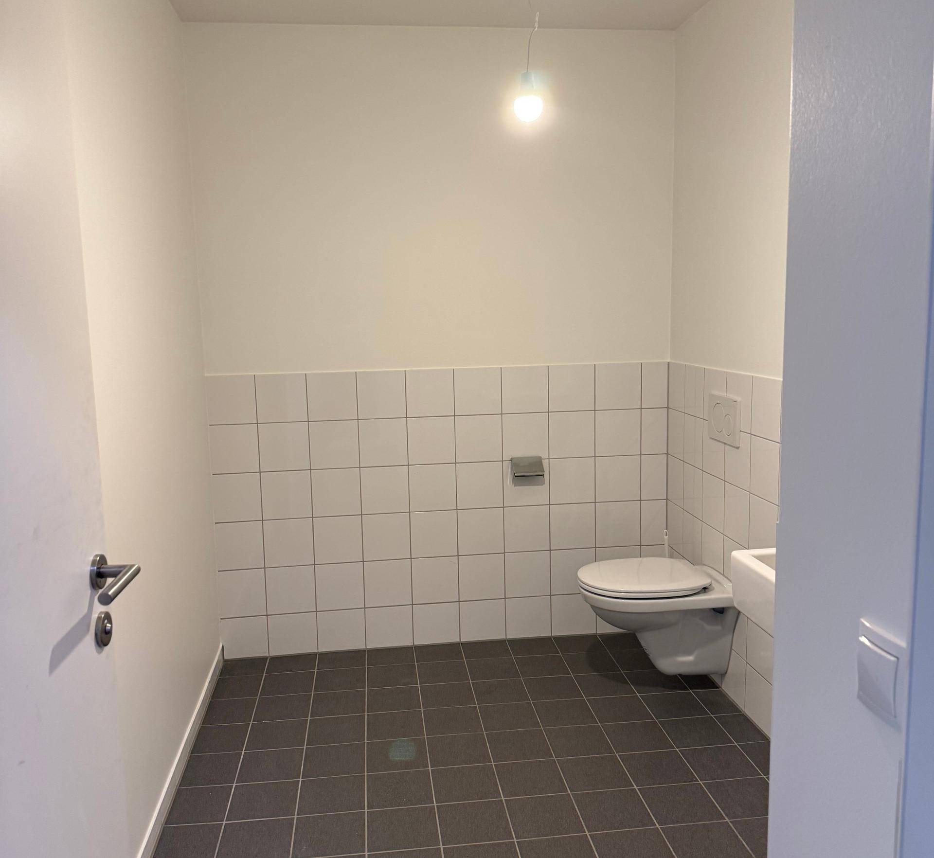 Saubere Gästetoilette mit weißen Fliesen an der Wand und dunklem Fliesenboden, ausgestattet mit Wand-WC und kleinem Waschbecken.
