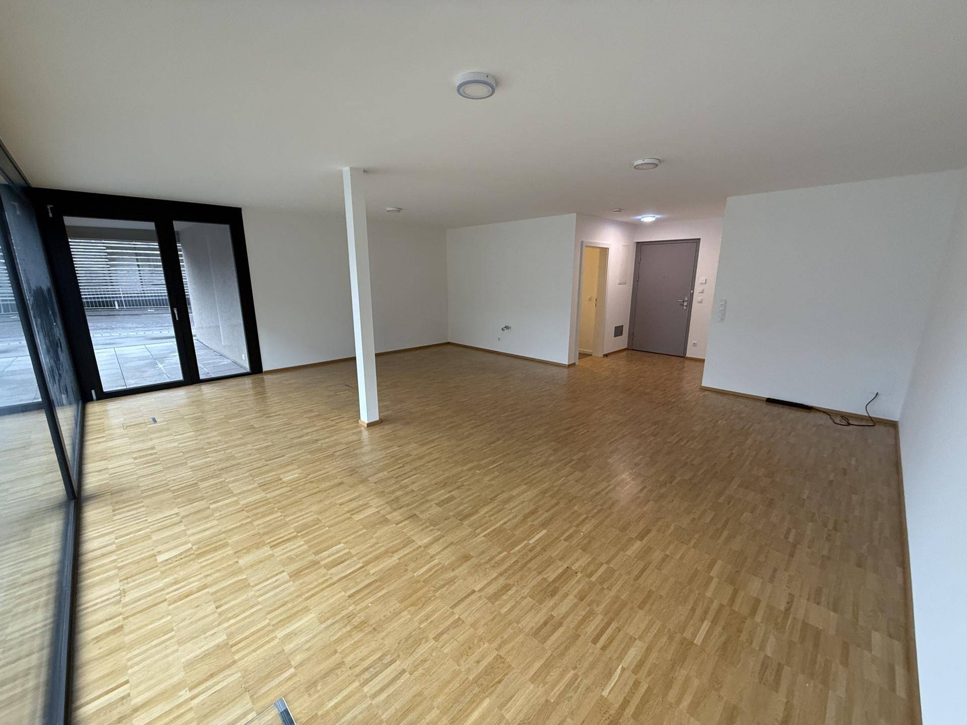 Geräumiger, unmöblierter Wohnbereich mit Holzboden, großen Fenstern und Zugang zu einem Außenbereich, ideal für neue Mieter.