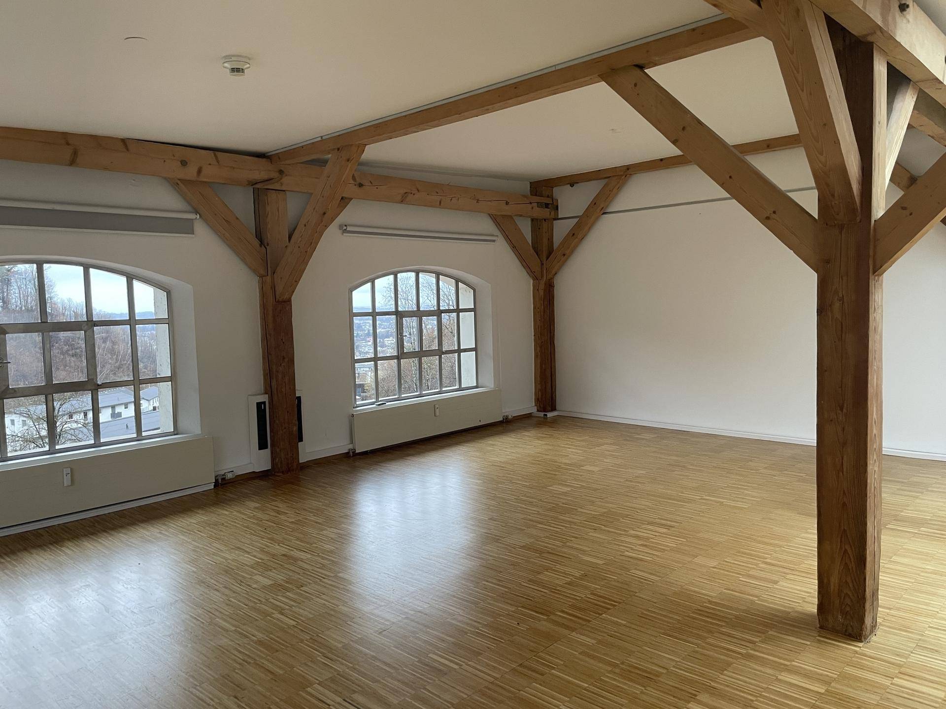 Geräumiger, leerer Raum mit Holzboden, sichtbaren Holzträgern und großen Bogenfenstern, die viel Licht hereinlassen.