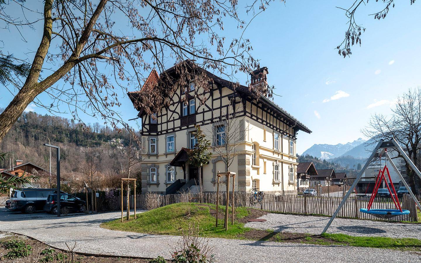 Historisches Gebäude mit Fachwerk und einem Spielplatz im Vordergrund, umgeben von Bergen und Bäumen.