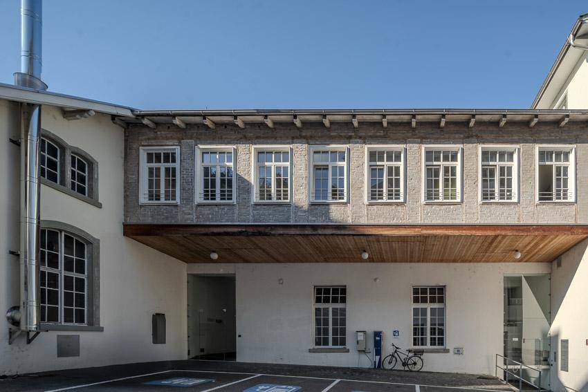 Außenansicht eines Gebäudes mit Parkplätzen, einer Ladestation für Elektrofahrzeuge und einer Fahrradabstellmöglichkeit.