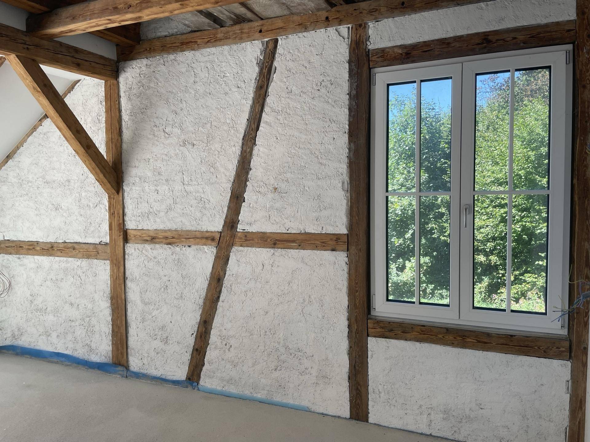 Innenwand mit sichtbarem Fachwerk und Putz, kombiniert mit einem modernen Fenster, das einen schönen Grünblick bietet.