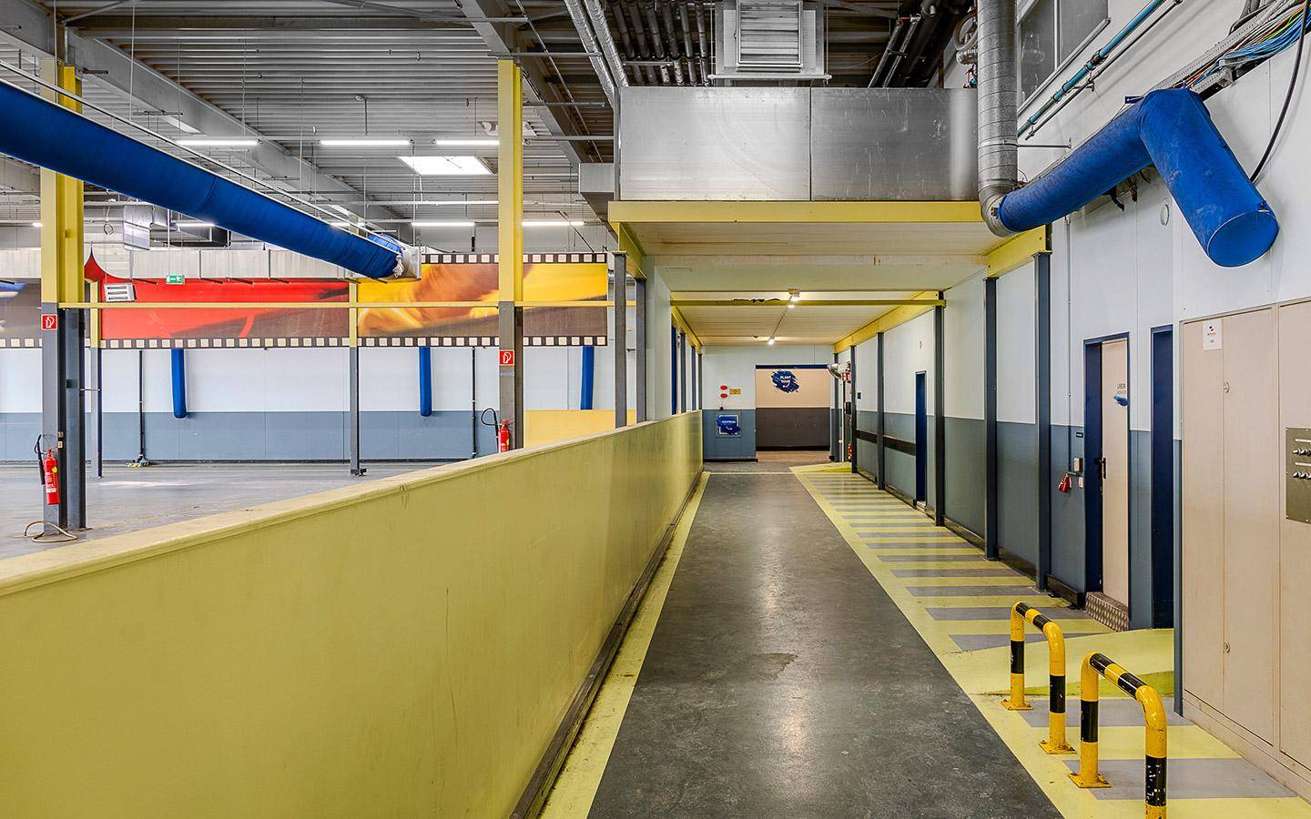 Industrieller Flur mit gelben Geländern, blauen Rohren und funktionalen Türen in einem Gewerbegebäude.
