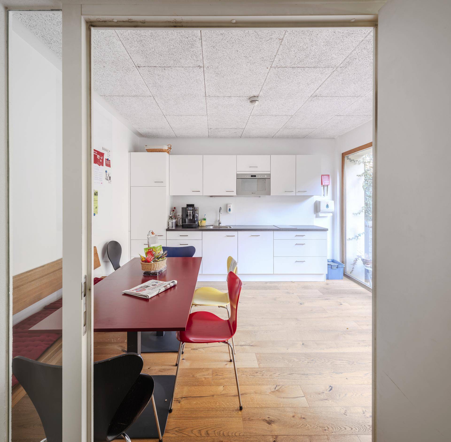 Moderne Küche mit weißen Schränken und einem Essbereich mit Holzboden, ausgestattet mit farbigen Stühlen und viel Tageslicht.