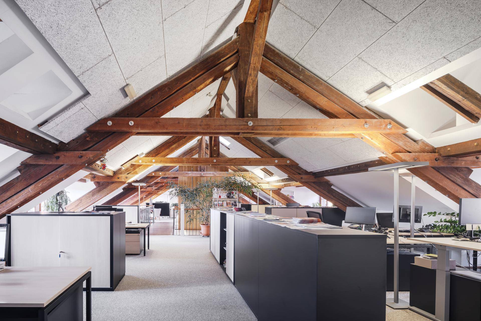 Großes offenes Büro im Dachgeschoss mit sichtbaren Holzbalken, hohen Decken und mehreren Arbeitsplätzen.