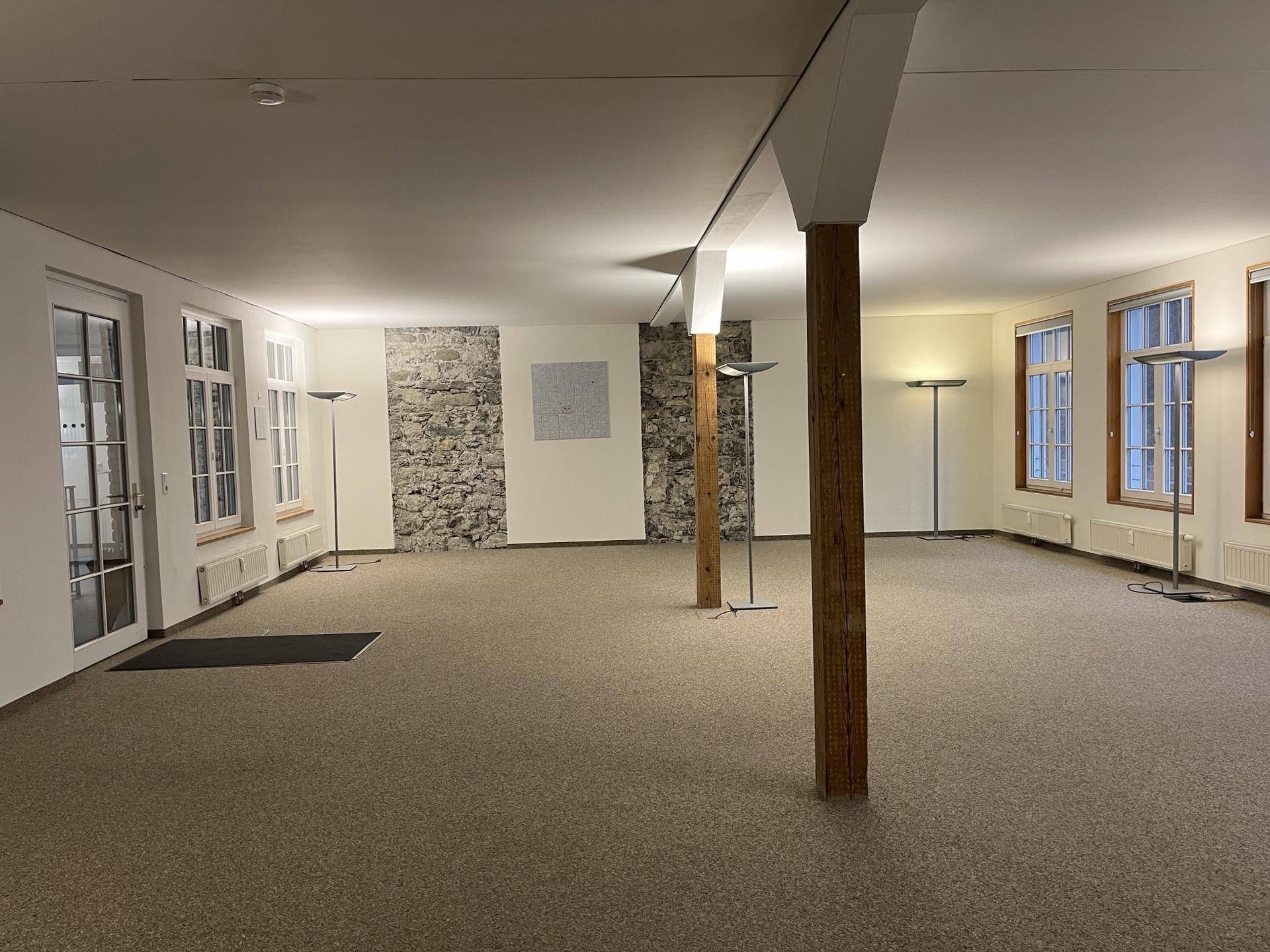 Geräumiger, leerer Raum mit Teppichboden, freigelegter Steinwand und rustikalen Holzstützen.