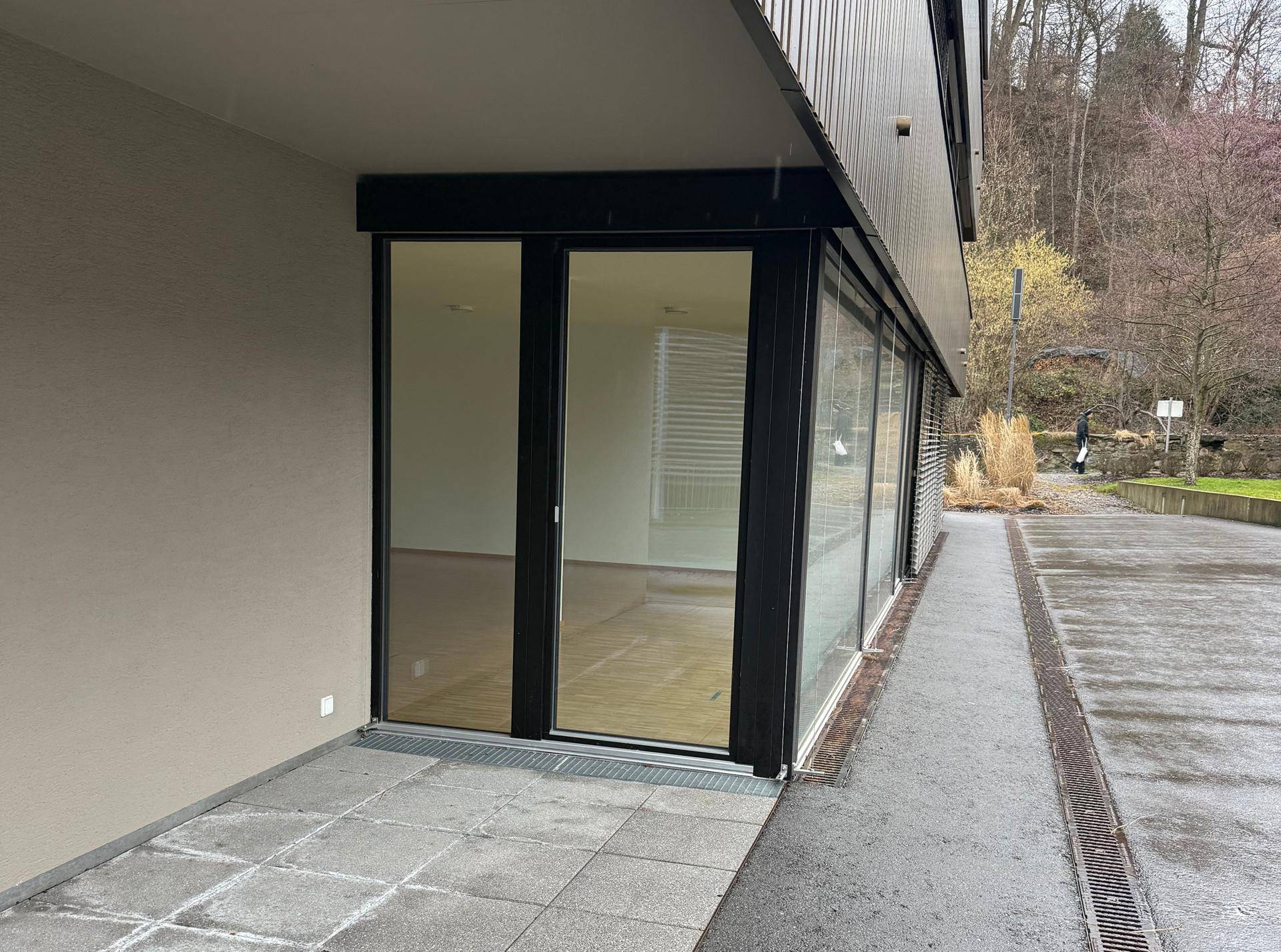 Eingangsbereich im Erdgeschoss mit großen Glasfronten und einem gepflasterten Außenbereich, umgeben von moderner Architektur.