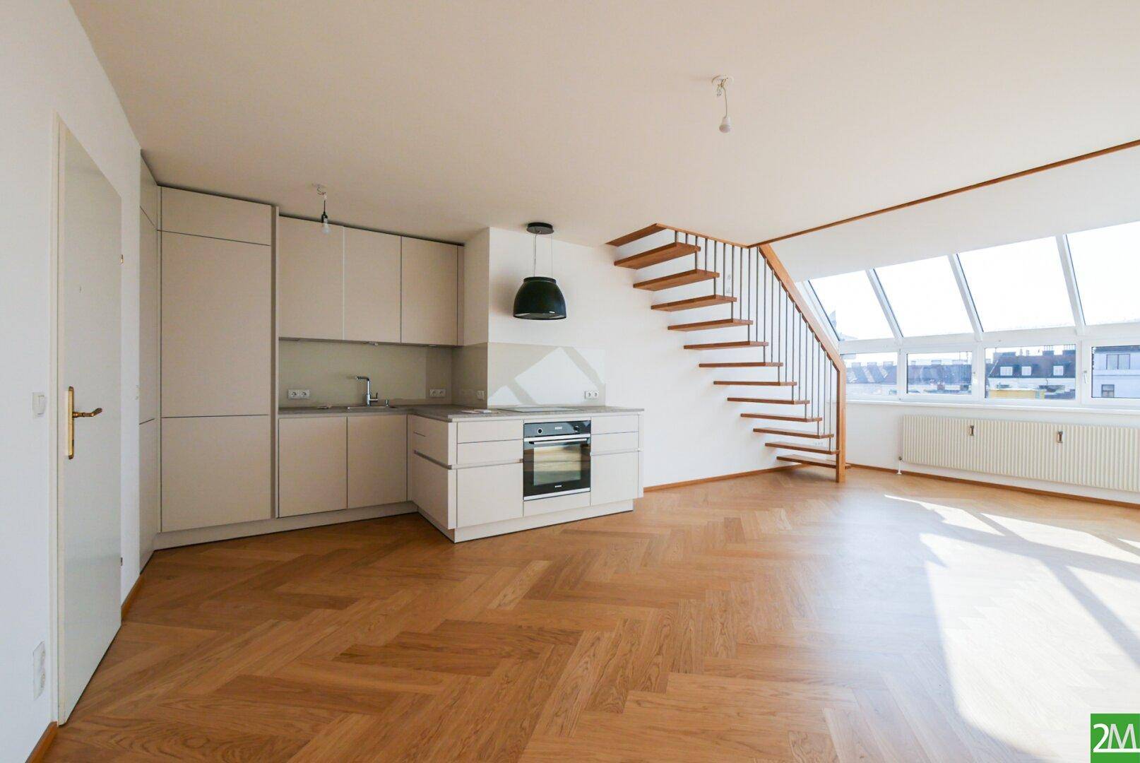 Helle Küche mit Einbaugeräten und Holzparkett, offen zu einem Wohnbereich mit großen Fenstern.