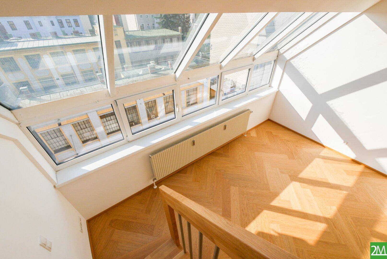 Blick von oben auf den hellen Wohnbereich mit Holzparkett und großen Fenstern, die viel Licht hereinlassen.