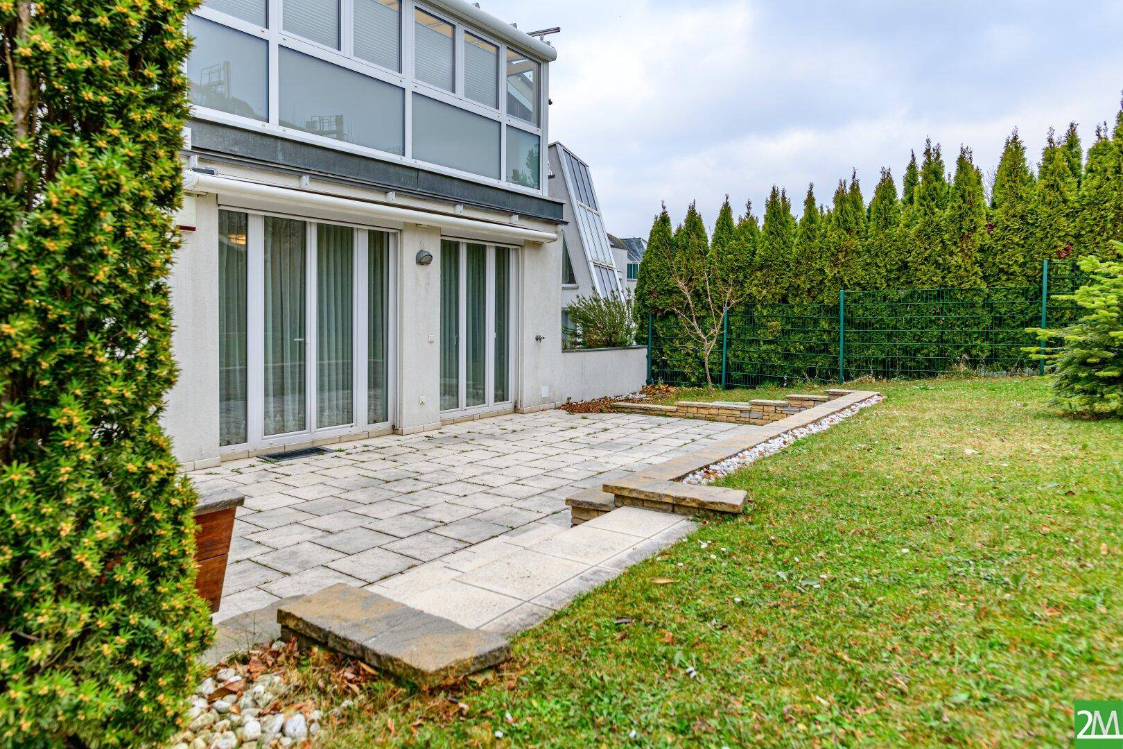 Gepflasterte Terrasse neben dem Haus mit angrenzender Rasenfläche und hohen Hecken.