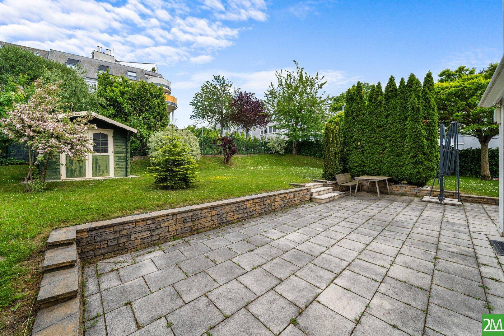 Geräumige gepflasterte Terrasse mit Sitzgelegenheiten, Rasen und einem kleinen Gartenhaus.