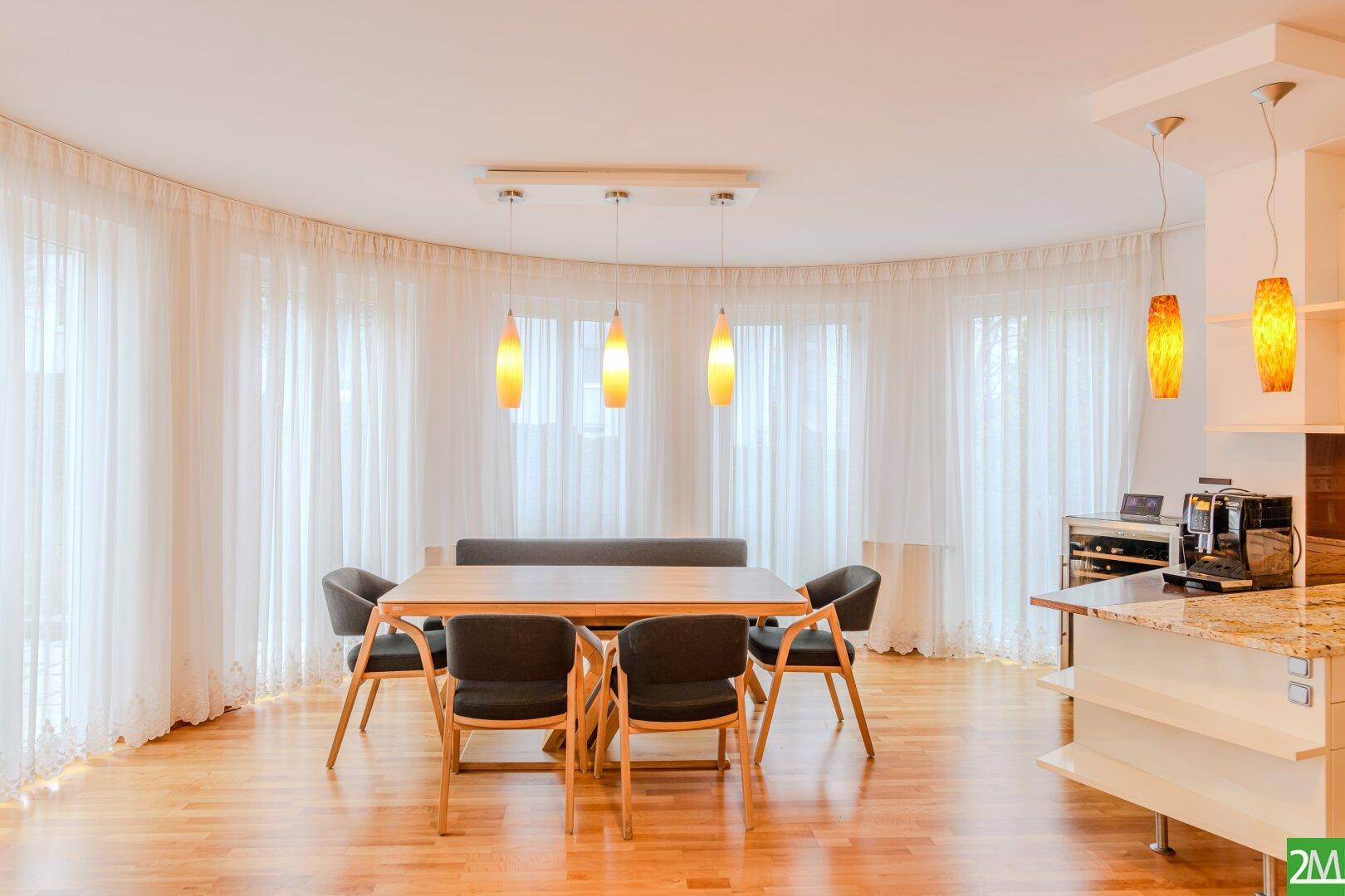 Heller Essbereich mit Holztisch, modernen Pendelleuchten und großen Fenstern mit Vorhängen.