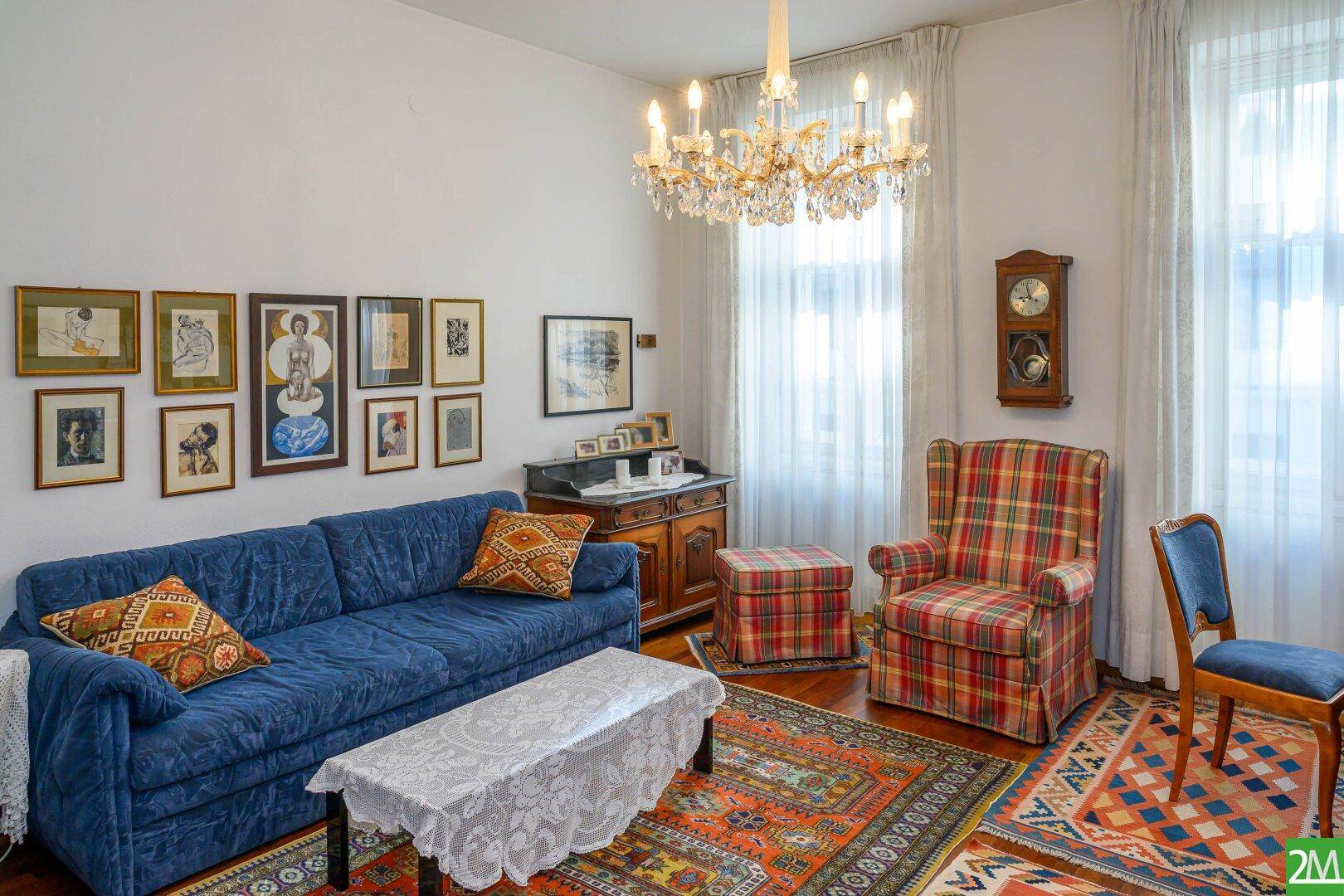 Klassisches Wohnzimmer mit blauem Sofa, kariertem Sessel, Kronleuchter und gemusterten Teppichen.
