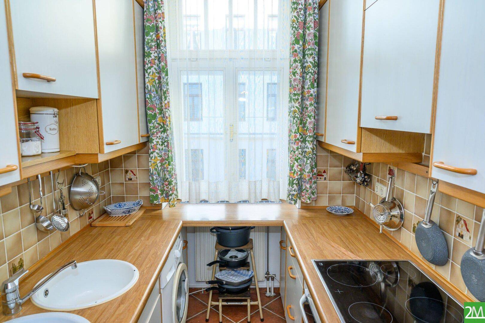 Kleine Küche mit weißen Schränken, Holzarbeitsplatten und einem Fenster mit Blumenmuster-Vorhängen.