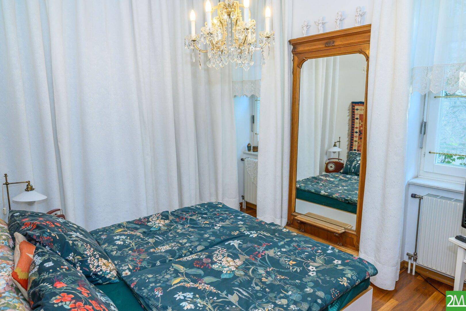 Gemütliches Schlafzimmer mit Doppelbett, großem Spiegel, Kronleuchter und floraler Bettwäsche.