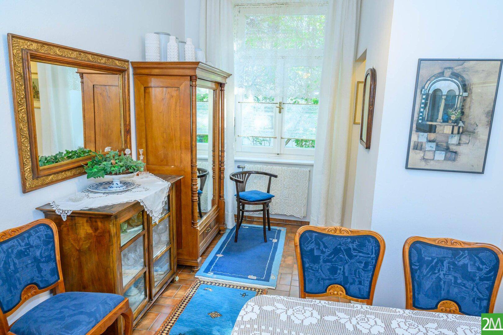 Traditioneller Essbereich mit antiken Holzmöbeln, blauen Stühlen und einem großen Fenster mit Vorhängen.