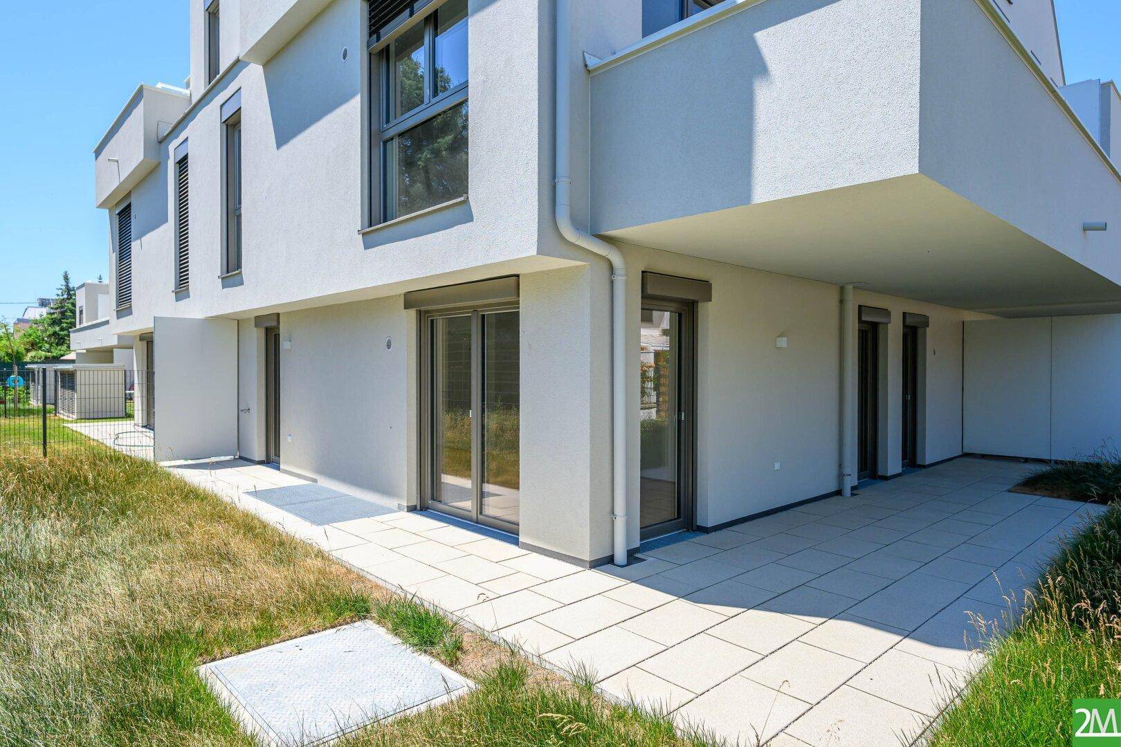 Moderne Gebäudefassade mit großen Fenstern und einer gepflasterten Terrasse im Erdgeschoss.