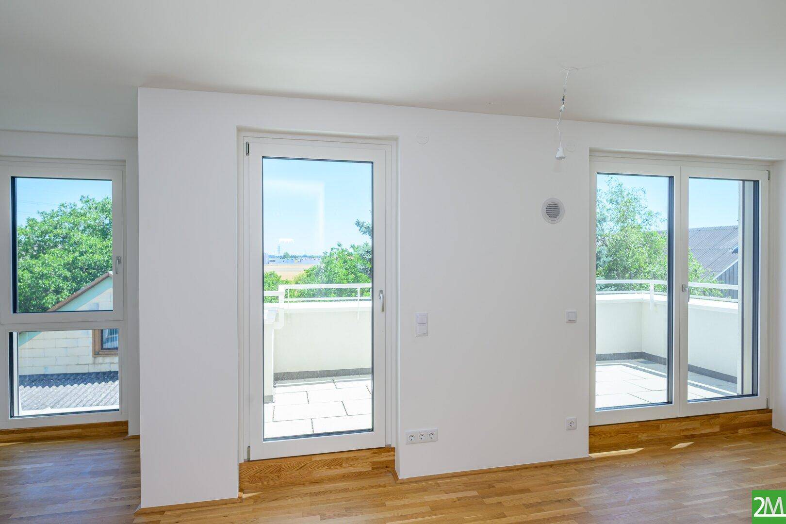 Lichtdurchfluteter Wohnraum mit Holzboden und mehreren Fenstern, die Zugang zum Balkon bieten.