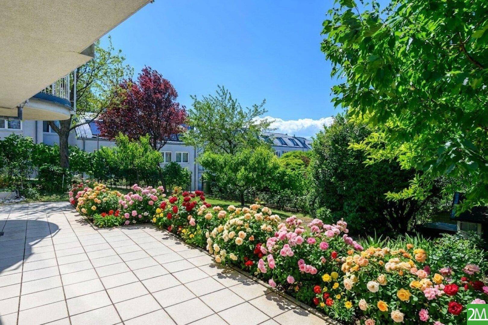 Geräumige Terrasse mit Fliesenboden, umgeben von bunten Blumenbeeten und viel Grün.