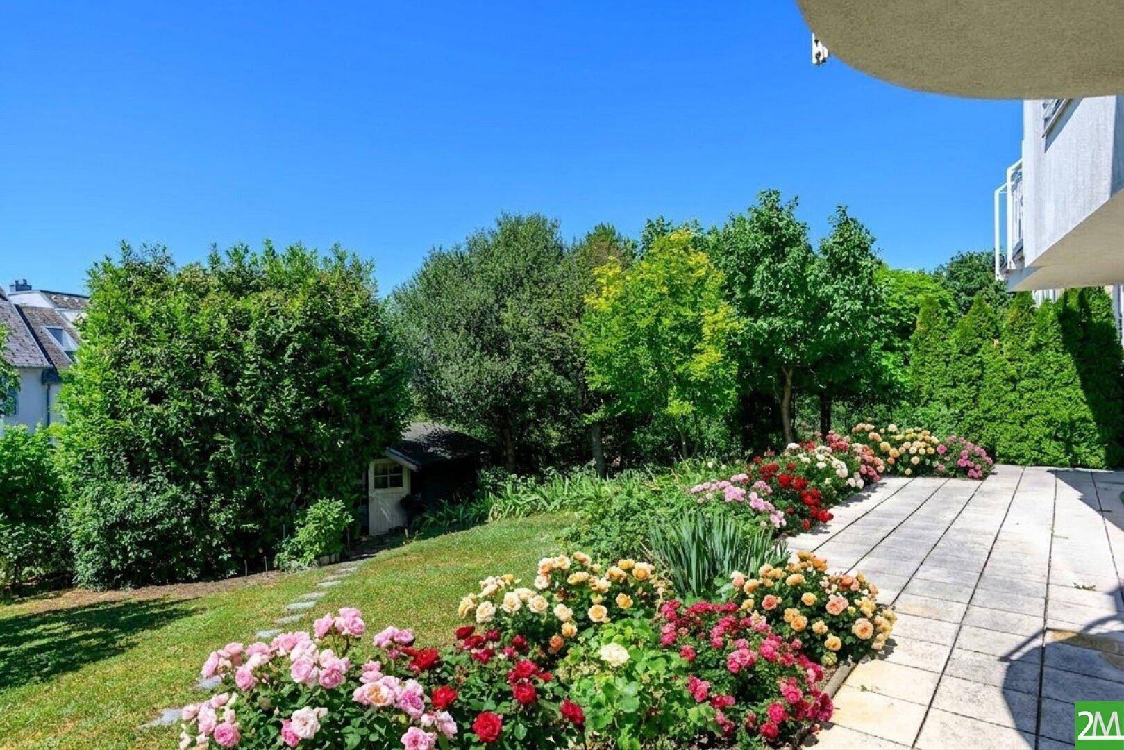Sonniger Garten mit Rasenfläche, Blumenbeeten und einem kleinen Gartenhaus im Hintergrund.