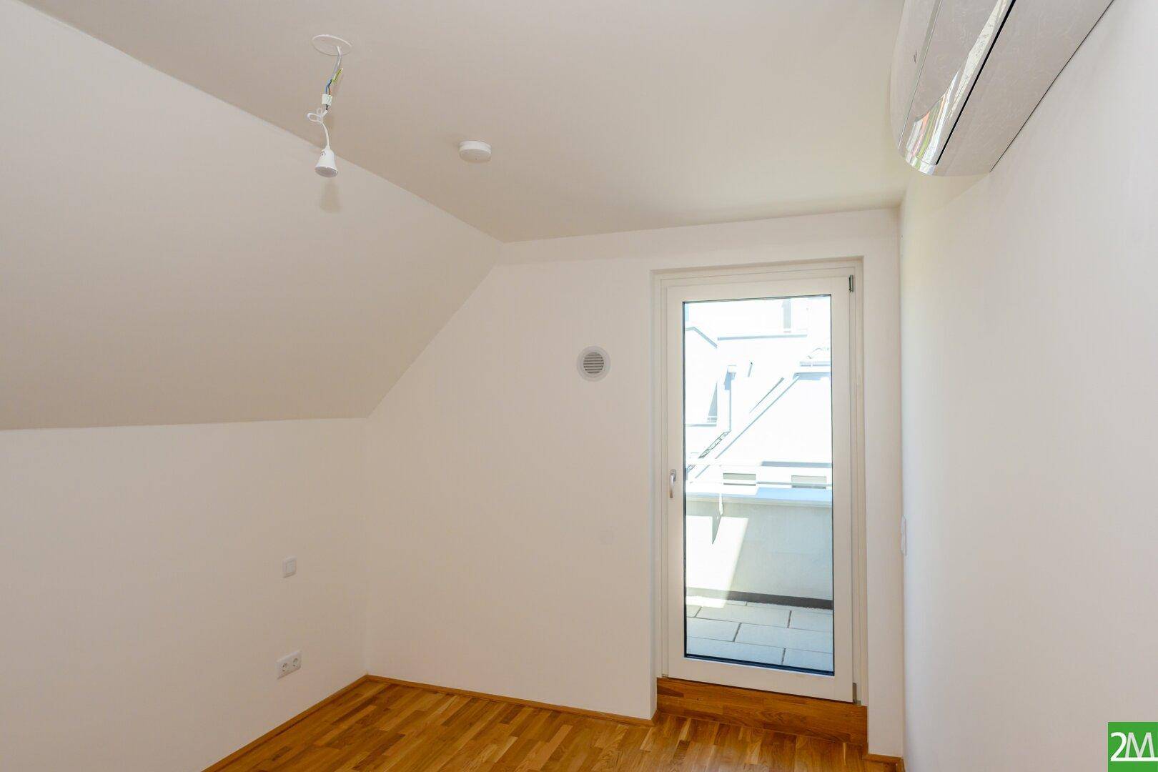 Leeres Zimmer mit Holzboden, Dachschräge, Klimaanlage und Zugang zu einem Außenbereich.