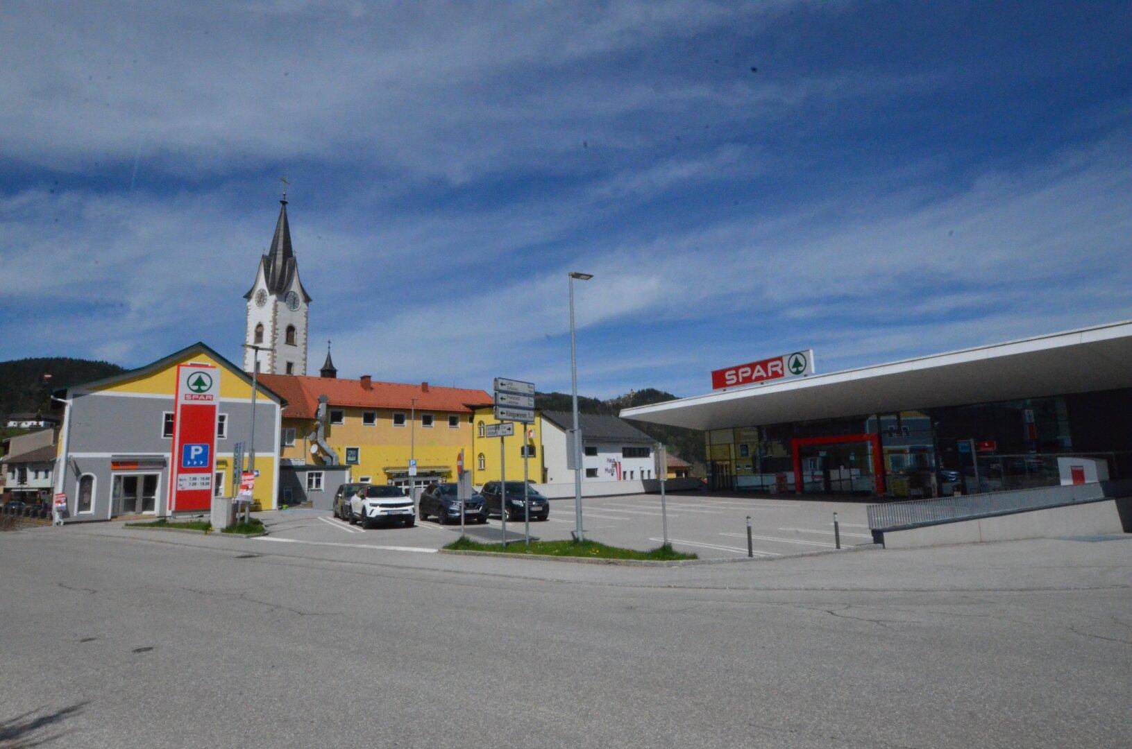 Weitläufige Außenansicht mit Supermarkt, Parkplätzen und einem Kirchturm unter blauem Himmel.