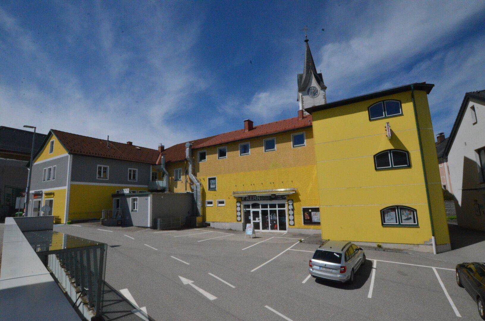Gelbes Gebäude mit Parkplatz und einem Kirchturm im Hintergrund unter einem hellblauen Himmel.