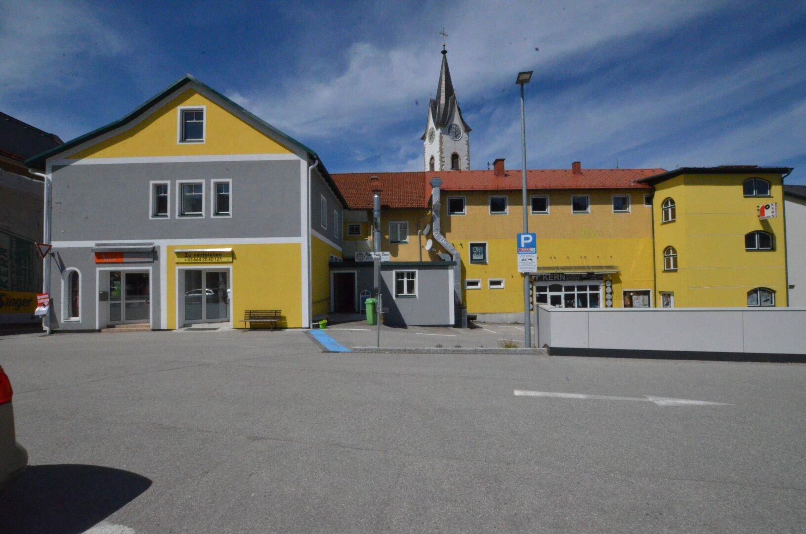 Außenansicht eines Gebäudes mit gelber und grauer Fassade und einem Kirchturm im Hintergrund.
