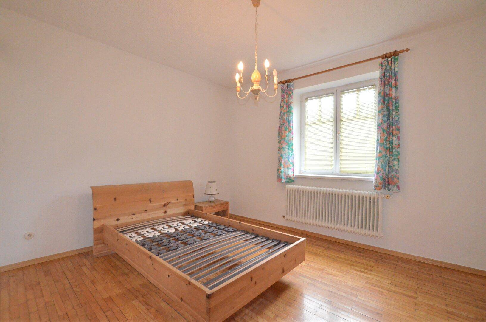 Gemütliches Schlafzimmer mit Holzbett, Parkettboden und einem Fenster mit Vorhängen.