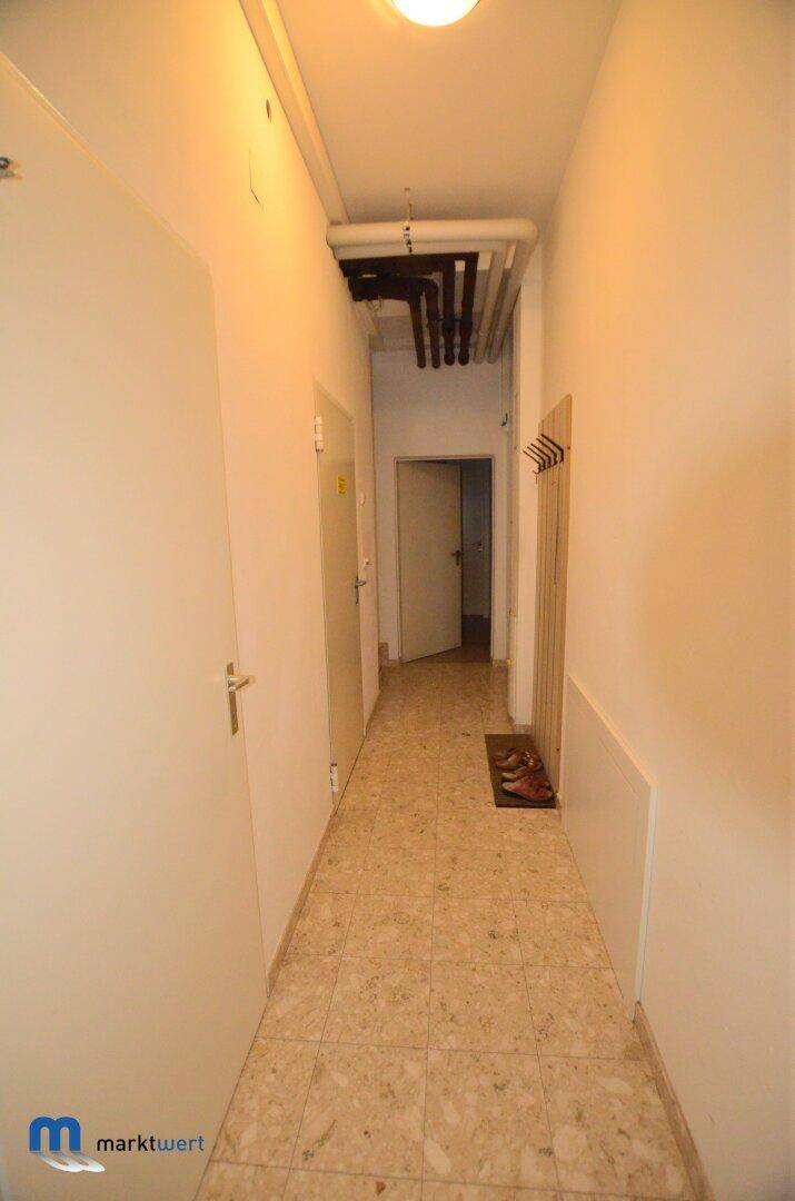 Langer, schmaler Flur mit Fliesenboden und sichtbaren Rohren an der Decke.