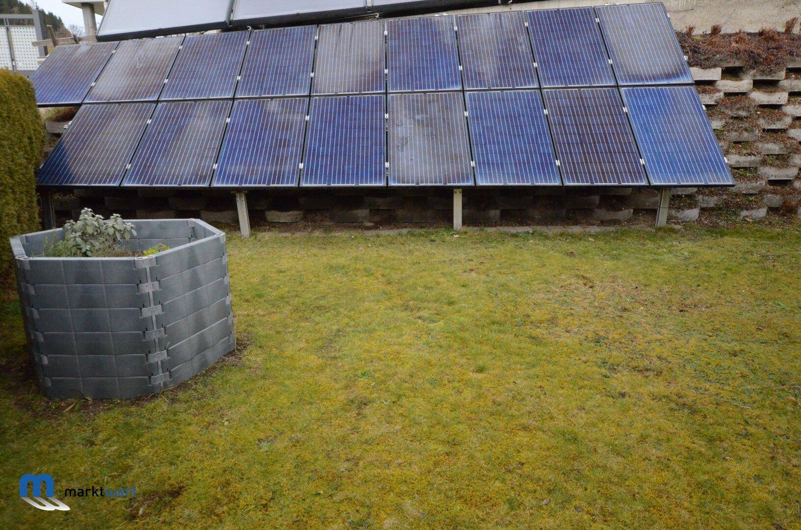 Gartenbereich mit Photovoltaikanlage und einem Hochbeet auf einer Rasenfläche.